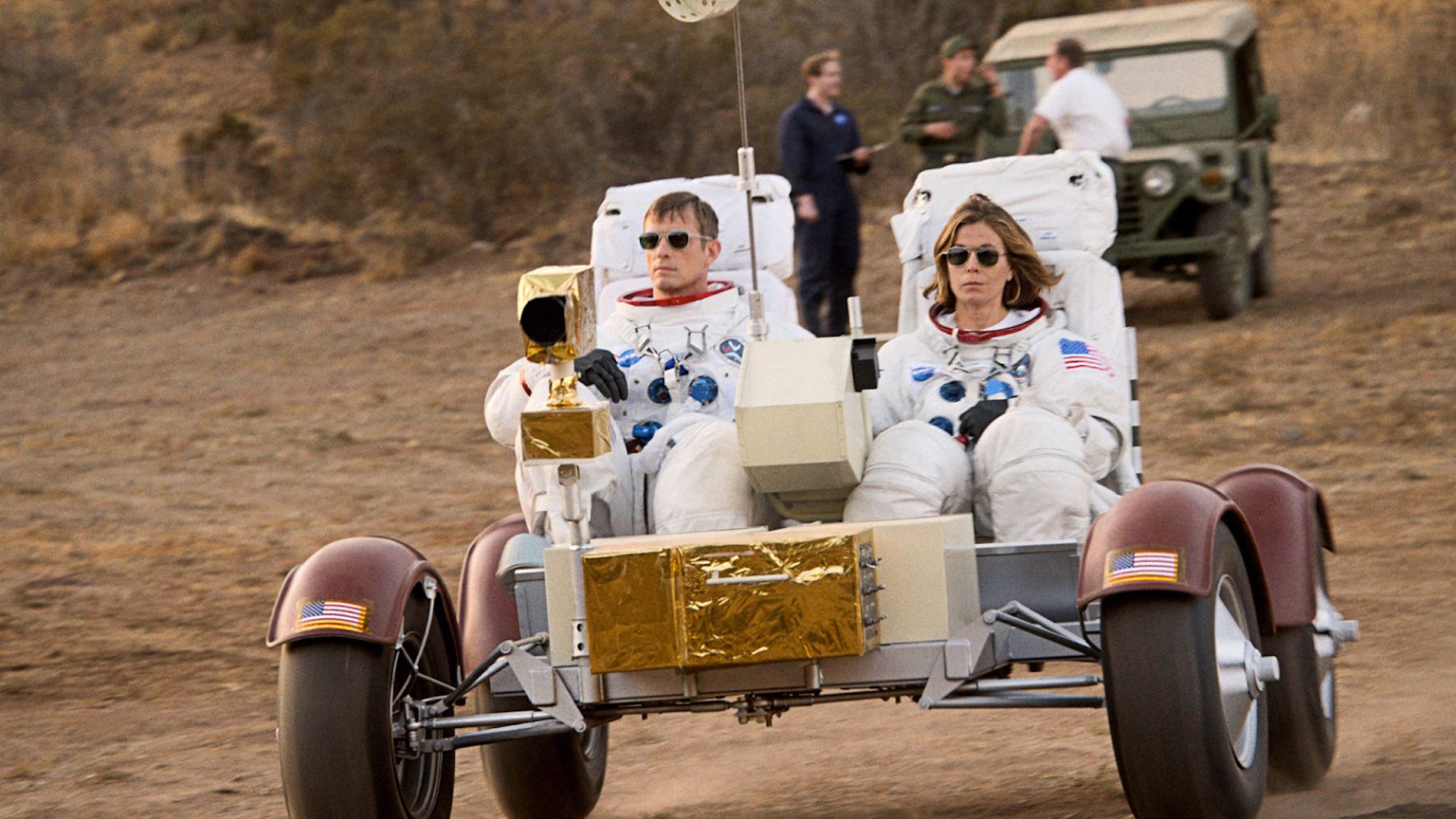 Two astronauts drive a buggy across dusty terrain. 