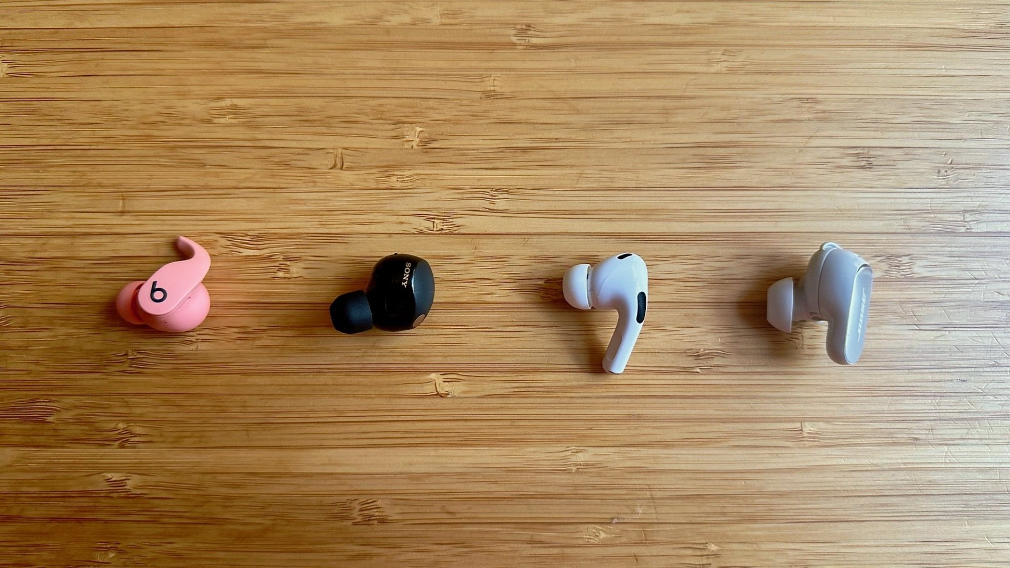 One earbud of the following on a wooden table: Beats Fit Pro, Sony WF-1000XM5, Apple AirPods Pro, and Bose QuietComfort Ultra.