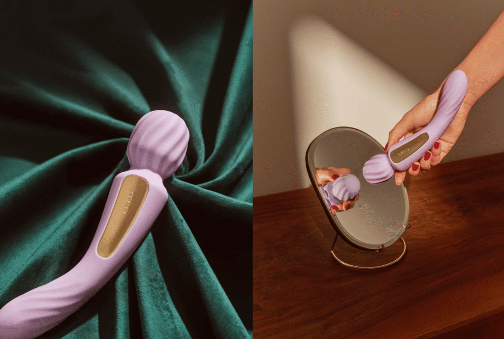 A person sits in a candlelit bath, their hand resting on a wooden bath caddy. On the caddy sits a lilac-colored, dual-ended LELO Switch vibrator next to a glass of wine and a book, creating a cozy and intimate scene.