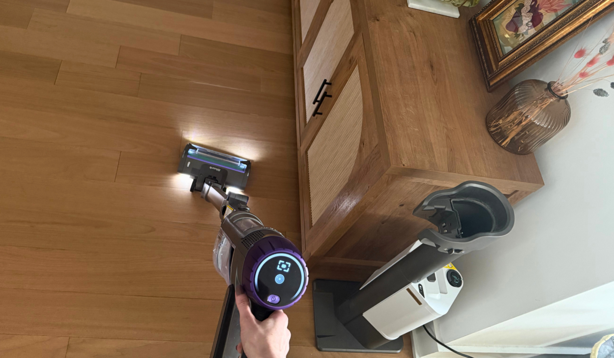 Person's hand cleaning hardwood floor with Shark cordless vacuum near auto-empty dock